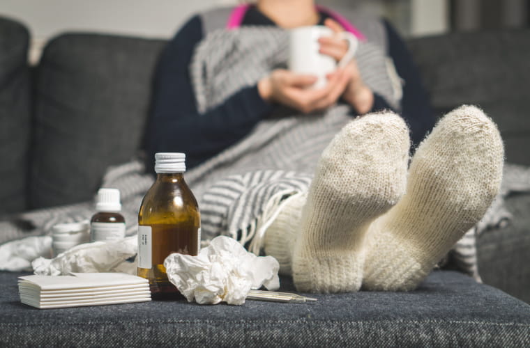 A sick woman is on the couch drinking a hot beverage to get well from the flu. She has cold medicine and dirty tissues on the table.