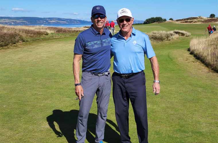 Two men on a golf course with blue water and mountains in the background. Blue sky is above.