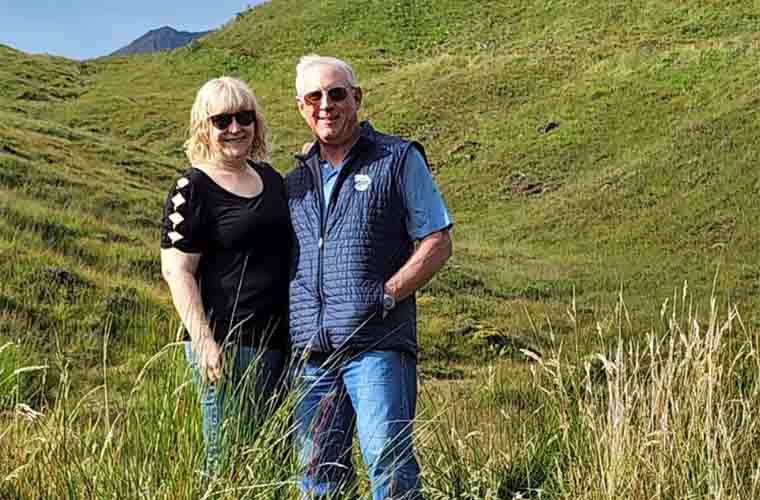 Man and woman standing outside in the green grassy hills with mountains behind them.