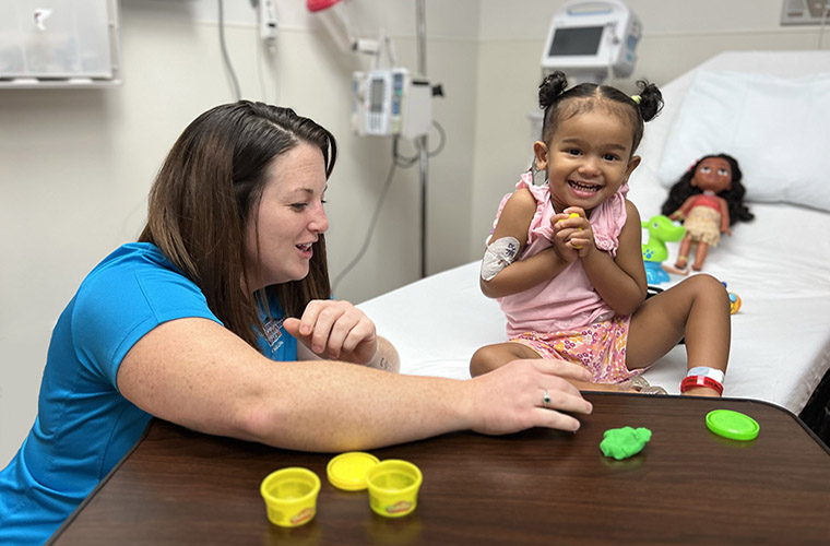 A female child life specialist entertains a young female patient who is smiling from her hospital bed.