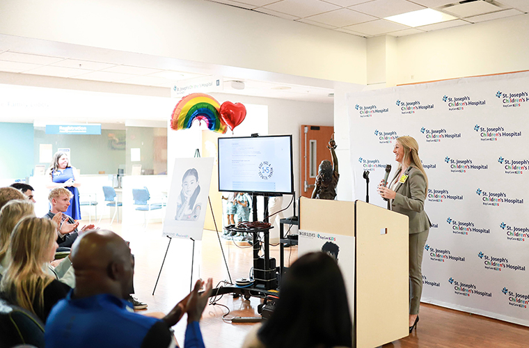 Dr. Christina Canody wearing a gray pantsuit stands behind a podium with a microphone addressing a group on seated attendees. She is flanked by a TV screen and a BayCare backdrop with the words St. Joseph's Children's Hospital and BayCare Kids is behind her. 