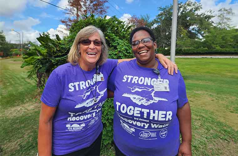Two women in purple T-shirts standing outside smiling at the camera.