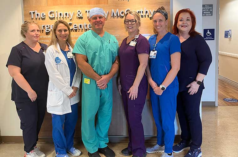 A group of people dressed in different colored hospital scrubs smile for a picture as they stand in front of a surgical center.