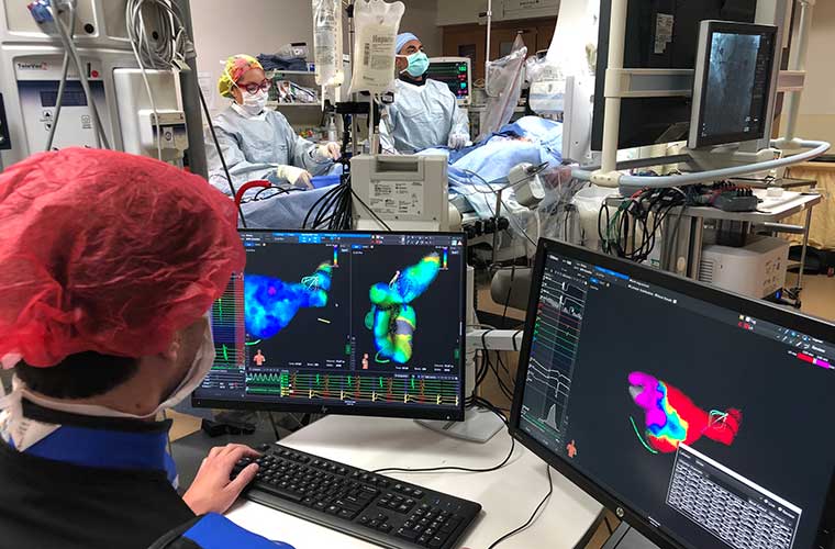 Two clinicians dressed in full white surgical gowns with a blue cap and rainbow tie dye cap respectively, perform the cardiac ablation looking at a big flat screen tv in front of them showing the inside of the heart to guide them with the patient laying on the operating table in front of them. There is a member of the surgical team with a red surgical cap watching the progress from 2 screens that show the inside of the heart with color coding's.