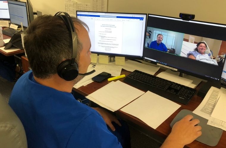 Virtual Nurse Dave Paliga is sitting behind a desk with papers and two computer screens in front of him chatting online with patient Samantha Anderson. The screen on the left shows discharge notes and the screen on the right shows his image and her image.