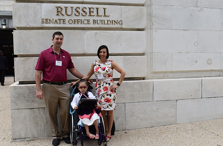A man and woman and child in a wheelchair are smiling and posing in front of a building in the nation's capitol. 