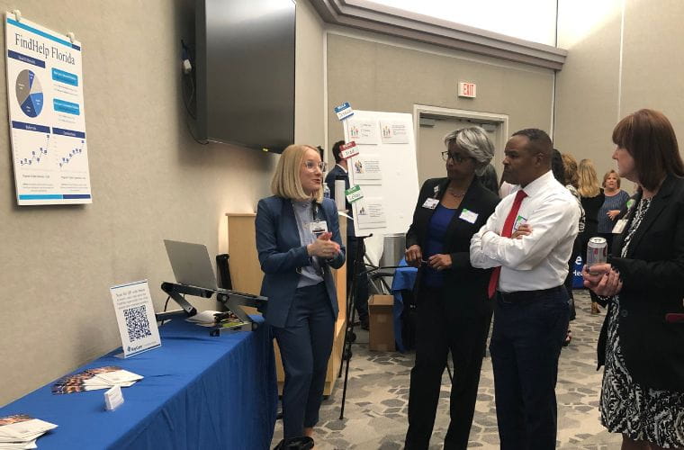 A young woman with short blonde hair wearing glasses and a navy suit speaks to two women and one man in a small ballroom setting. A table with a blue tablecloth is to the left with brochures and a laptop. There is a sign on the wall with a pie graph and the words FindHelp Florida. Other attendees are in the background.