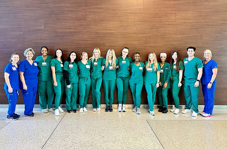 Men and women wearing blue and teal scrubs are standing in front of a wall and smiling. 