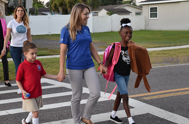 Woman holding hands with two children as they cross the street using a crosswalk. All three are smiling in the photo.