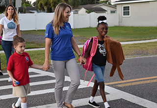 Woman holding hands with two children as they cross the street using a crosswalk. All three are smiling in the photo.