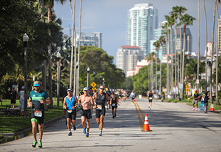 People in athletic gear run down a sunny street with palm trees and high-rise buildings in the background during an athletic event.  Caption: Athletes run through the streets of St. Petersburg during the 2023 St. Anthony’s Triathlon. Registration will open for the 2024 event on Monday, Oct. 2.