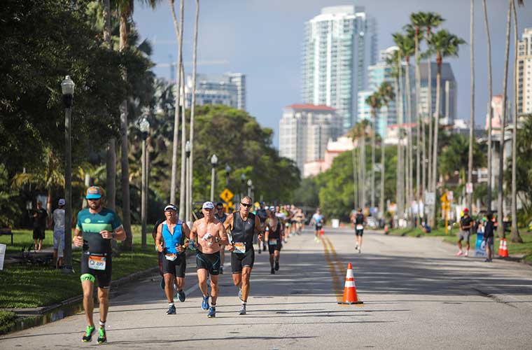 People in athletic gear run down a sunny street with palm trees and high-rise buildings in the background during an athletic event.  Caption: Athletes run through the streets of St. Petersburg during the 2023 St. Anthony’s Triathlon. Registration will open for the 2024 event on Monday, Oct. 2.
