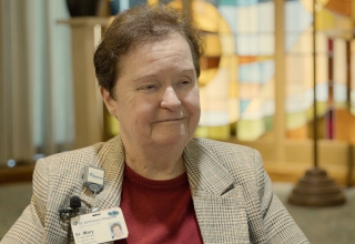 A smiling headshot of Sister Mary McNally wearing a maroon shirt and brown plaid jacket.  She has short brown wavy hair and is wearing a name badge on the right side of her jacket. The hospital lobby is in the background. 