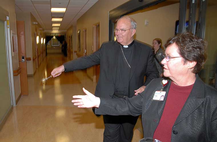 Sister Mary McNally and Bishop Emeritus Robert Lynch motion toward a wall at St. Anthony's Hospital. She is wearing a marron shirt, a black jacket and glasses. He is wearing dark clergy attire and glasses. 