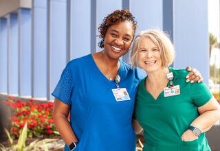 A middle-aged dark-skinned woman in blue scrubs and an older white skinned woman in green scrubs smiling and hugging.