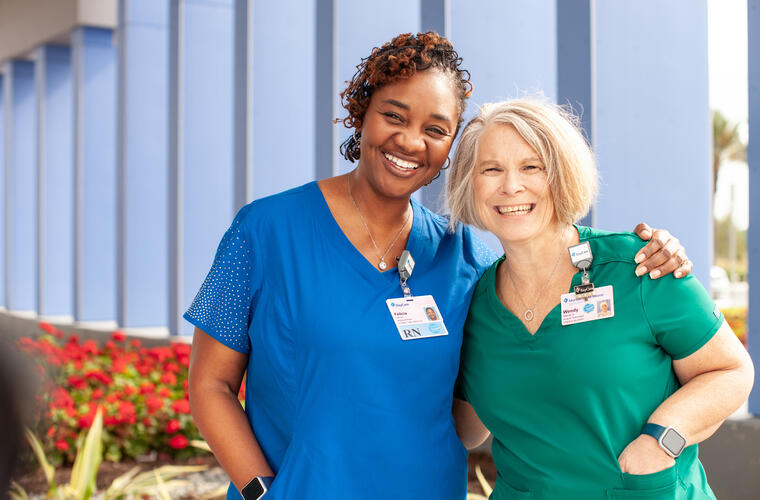 A middle-aged dark-skinned woman in blue scrubs and an older white skinned woman in green scrubs smiling and hugging.