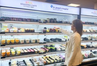 A woman with short blonde hair wearing a white blazer reaches for a food item in a cafeteria. There is signage that reads SEASONS CAFE technology by amazon and St. Joseph's Hospital BayCare Health System above the cooler.