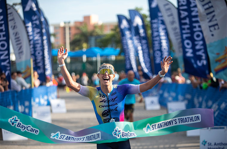 A woman dressed in athletic clothing smiles broadly while holding the finish line tap after winning a race.