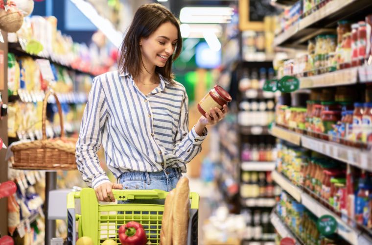 A young woman is smiling looking down at a jar of salsa reading the nutrition label. She has shoulder-length brown hair, is wearing a blue and white striped shirt and jeans and standing in the aisle of a grocery store. She is pushing a lime green cart with groceries inside including two baguettes and a red pepper.