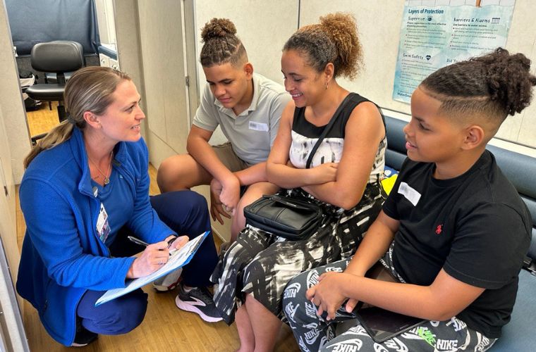 Nurse in blue scrubs holding a clipboard interviews a mother and two sons inside a mobile medical clinic.