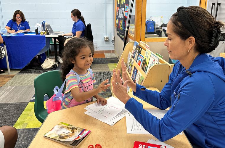 Female educator sits at table with preschool-aged girl holding up ten fingers to practice counting in a developmental screening.