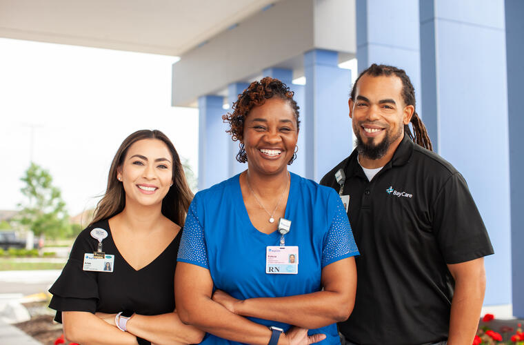 A diverse group of three BayCare team members, two women and one man, smiling and standing side by side in front of a BayCare facility wearing different work attire and BayCare badges.