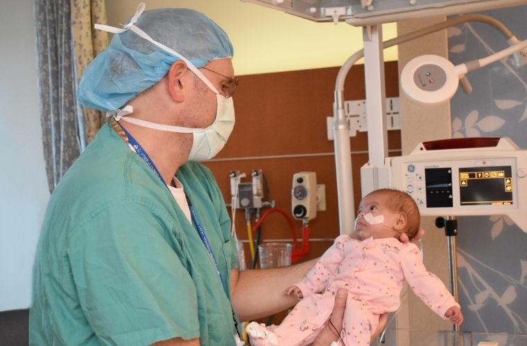  A doctor in a hospital gown and mask standing in room with medical equipment cradles a baby in his arms.