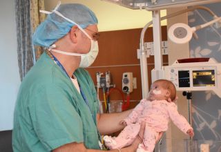  A doctor in a hospital gown and mask standing in room with medical equipment cradles a baby in his arms.