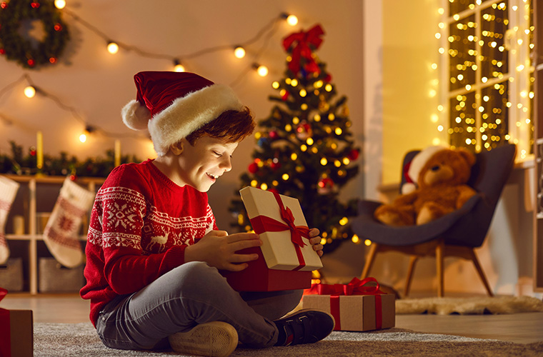 A young boy wearing a red sweater and a Santa hat sits on floor in front of a tree and opens a present.