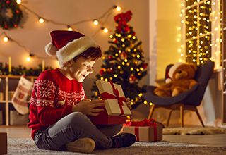 A young boy wearing a red sweater and a Santa hat sits on floor in front of a tree and opens a present.