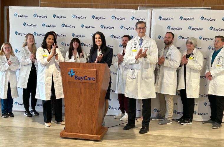 BayCare President and CEO Stephanie Conners stands front center behind a podium, with Dr. Sowmya Viswanathan to her left and Dr. Paul Lewis to her right in front of representatives of the residency program at a press conference on Feb. 1. The residents are wearing white lab coats, and everyone is standing in front of a BayCare backdrop.