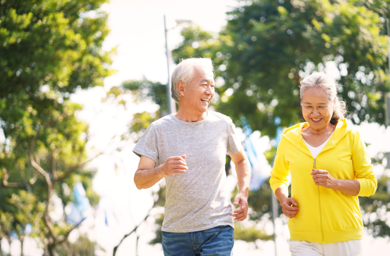A man and woman are smiling at each other as they jog outdoors and enjoy the sunny weather.