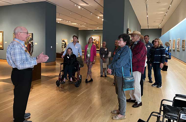 An older man dressed in a plaid shirt and dark slacks faces a group and gestures with his arms at a museum.