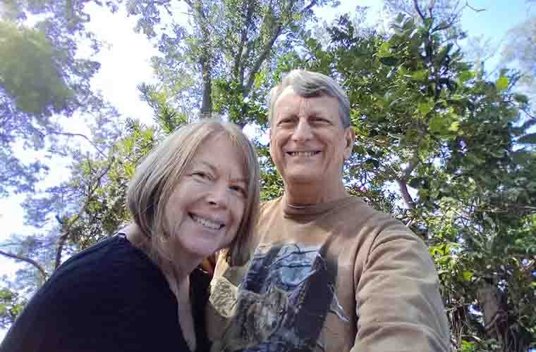A man and woman in their 60's smile at the camera. They are outside with trees and sky as the background.