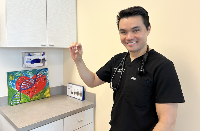 A man with black hair and wearing black scrubs is holding a small medical device and smiling at camera.