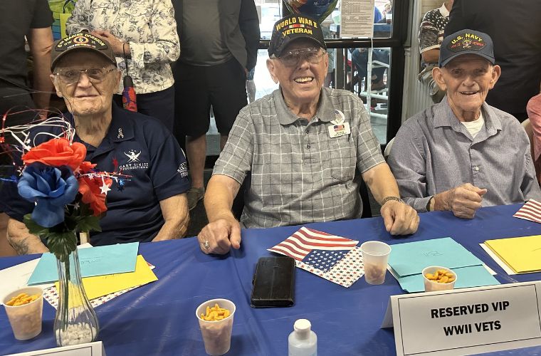 Three World War II veterans are seated at a table labeled "RESERVED VIP WWII VETS" during a community event, surrounded by people, with American flags and snacks on the table.