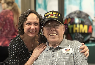 Two people smiling at the camera, one wearing a "World War II Veteran" cap and a badge that reads "Ray," and the other with curly hair, wearing a polka dot shirt.