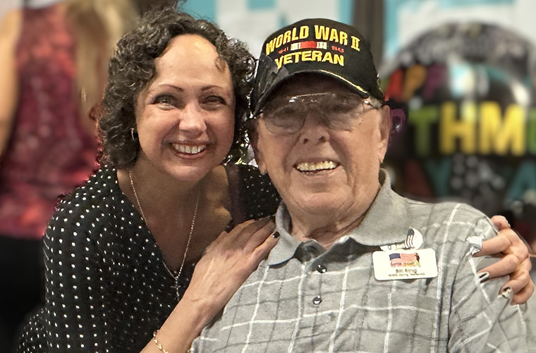 Two people smiling at the camera, one wearing a "World War II Veteran" cap and a badge that reads "Ray," and the other with curly hair, wearing a polka dot shirt.