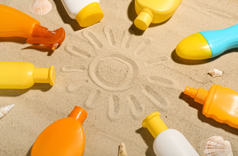 A variety of sunscreen bottles and lotion are arranged in a circle on the beach with a sun drawn in the sand.