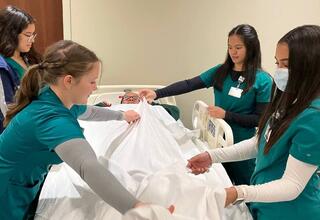 Four students in green hospital scrubs are helping a mock patient in a hospital bed by carefully adjusting the bed sheet as they smile kindly at the patient.