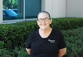 A middle-aged woman smiles for a photo outdoors while wearing black scrubs with the words "BayCare Laboratories" stitched on them. The woman is standing in front of some shrubs, outside of an office building. 