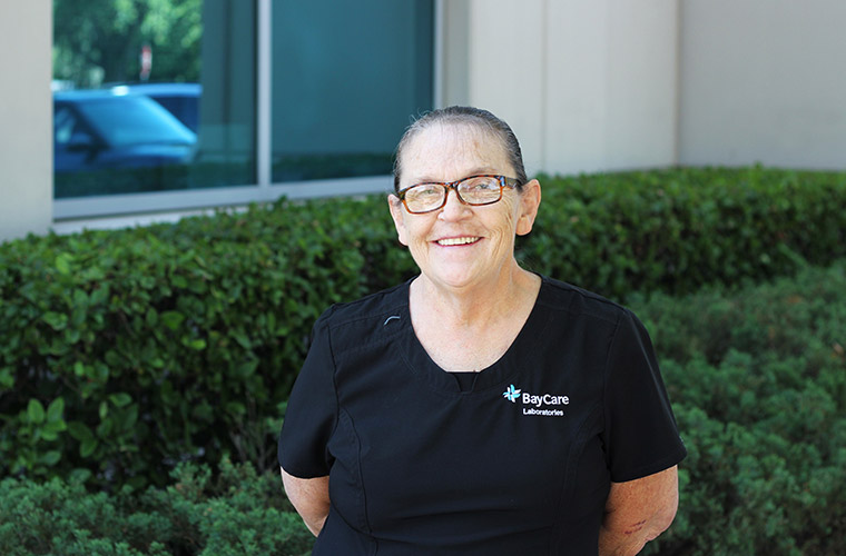 A middle-aged woman smiles for a photo outdoors while wearing black scrubs with the words "BayCare Laboratories" stitched on them. The woman is standing in front of some shrubs, outside of an office building. 