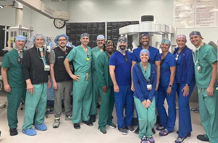 Twelve people in scrubs and surgical hats stand in a surgical suite and smile at the camera.