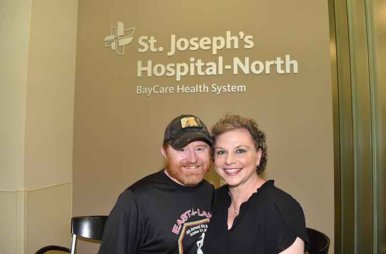 Liz, right, and her husband Dan at St. Joseph's-Hospital-North. "St. Joseph's Hospital-North, BayCare Health System" and BayCare logo are on the wall behind them. They are cheek-to-cheek.