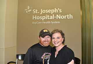Liz, right, and her husband Dan at St. Joseph's Hospital-North in June 2024. "St. Joseph's Hospital-North, BayCare Health System" and the BayCare logo are on the wall behind them. They are cheek-to-cheek.