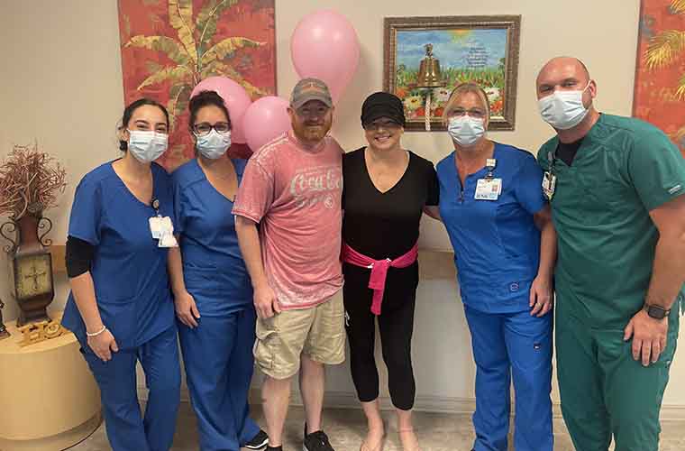 Three team members, two females and a male, in front of the bell in the infusion center lobby. There are also pink balloons with them and artwork on the wall. 