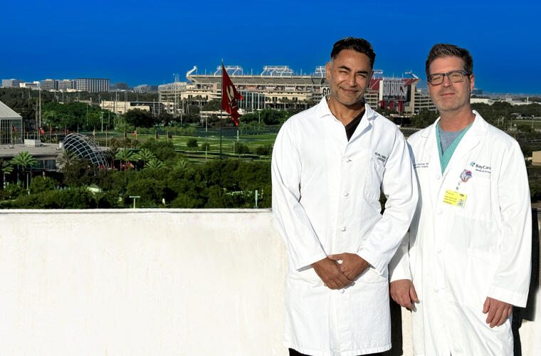 Two men in white lab coats stand on a rooftop with a football stadium behind them.