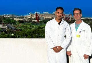 Two men in white lab coats stand on a rooftop with a football stadium behind them.