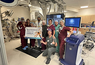 Ten hospital team members in scrubs are standing in an operating room holding a sign that says, "Congratulations! First case with FARAPULSE."