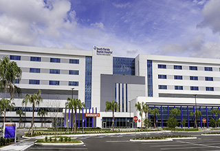 The exterior of South Florida Baptist Hospital. It’s a large hospital building against a blue sky with white clouds. Palm trees line the front of the building.